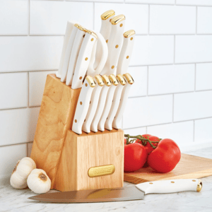 Knife block set with white handles and gold accents; includes various knives on a wooden block. In the background are tomatoes on a cutting board and garlic, placed on a marble countertop against a white tiled wall.
