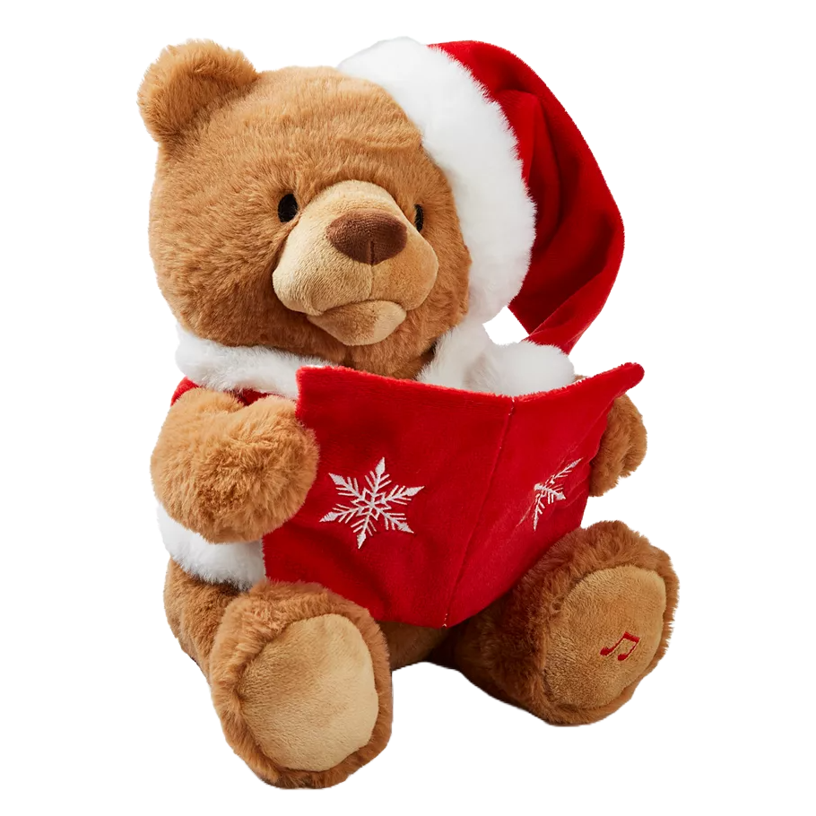 A plush bear wearing a Santa hat and red coat holds a red book with snowflake designs. It appears to be an animated toy designed for storytelling, featuring a music note emblem on its paw.