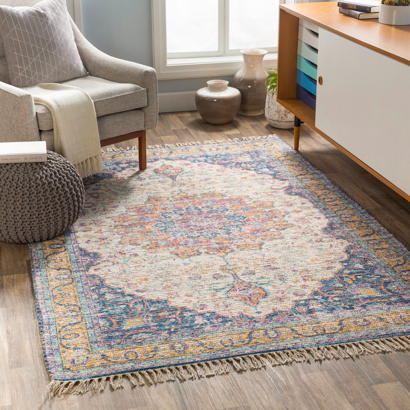 A 5' x 7' area rug with an intricate floral medallion design in multicolored hues, bordered by a blue and yellow pattern with fringed edges, placed on a wooden floor in a cozy, modern living space.