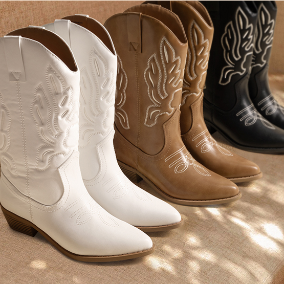 A collection of cowboy boots in white, brown, and black with detailed stitching, displayed on a cushioned bench in a room with wooden flooring.