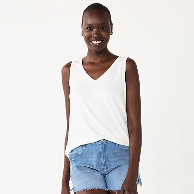 A smiling woman is wearing a white sleeveless V-neck tank top and denim shorts.