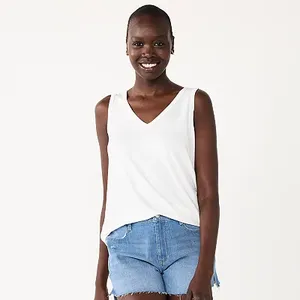 A smiling woman is wearing a white sleeveless V-neck tank top and denim shorts.