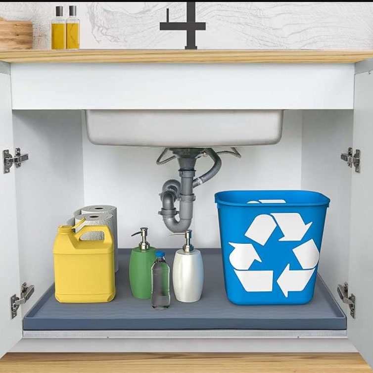Under-sink organization includes a blue recycling bin, yellow jug, roll of paper towels, and various bottles of soap and cleaning liquids, arranged on a protective tray with plumbing visible above.