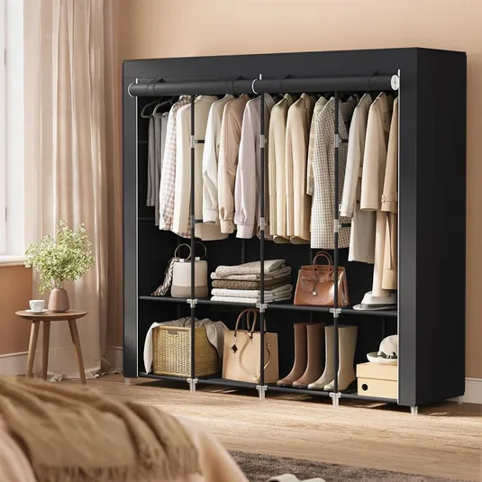 A black wardrobe contains beige and neutral-colored clothing on hangers, folded textiles, handbags, a wicker basket, and a pair of boots. A small table with a vase and cup is nearby, beside a curtain-draped window.