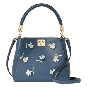 A blue Kate Spade crossbody bag featuring floral appliques with gold-tone hardware, a top handle, and a detachable strap, along with a central turn-lock closure.