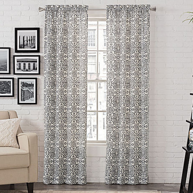 Black and white patterned curtains hang in a living room with a white brick wall background, accompanied by framed black-and-white photographs and part of a beige sofa.