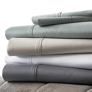 A stack of folded bedsheets in various colors, including gray, beige, white, and light blue, with decorative stitching near the edges.