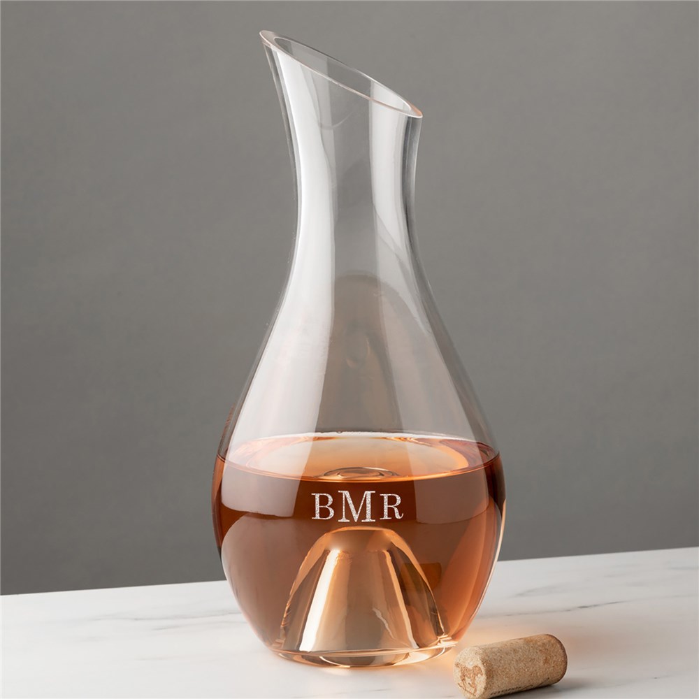A clear glass wine carafe with a slanted rim, engraved with the monogram \“BMR,\“ partially filled with rose-colored wine, sits on a white marble surface with a cork beside it.