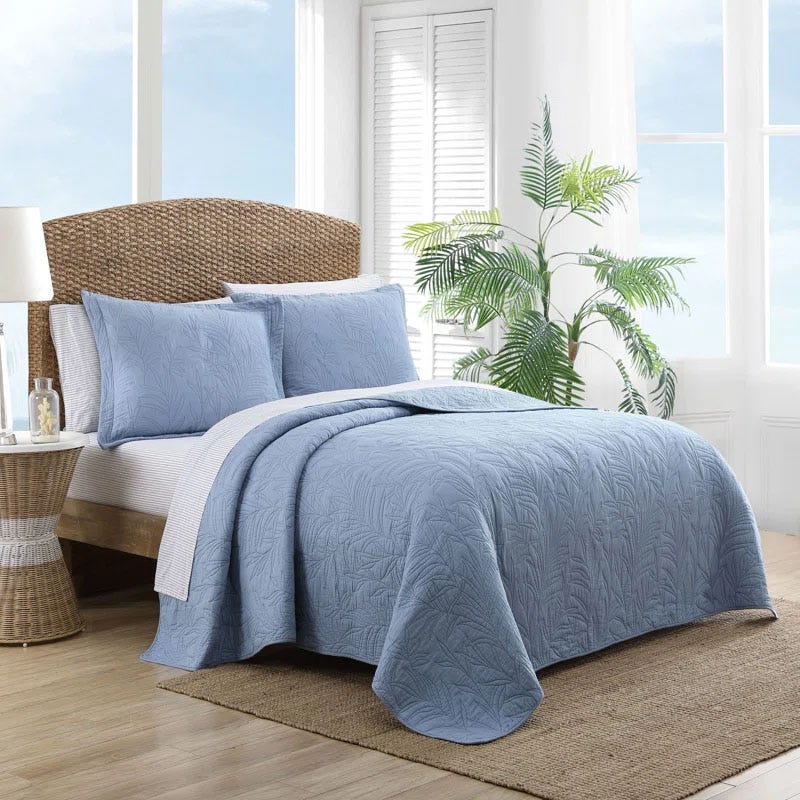 A cozy bedroom with a Tommy Bahama quilt set featuring a light blue quilt and matching shams with a leaf-patterned texture. The bed has a rattan headboard, and the room includes a potted plant and natural light from the windows.
