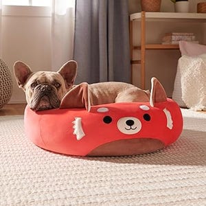 A French Bulldog is lying in a red panda-themed pet bed with plush ears and a cheerful face design. The bed is circular with a soft red exterior.