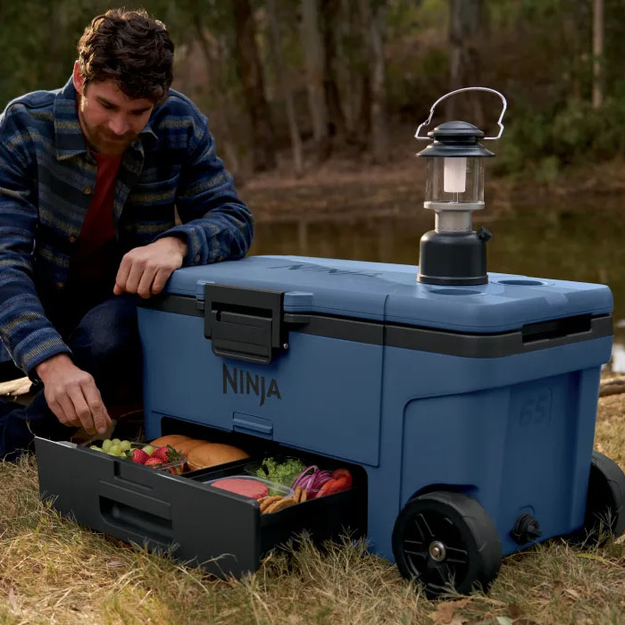 A blue Ninja FrostVault Wheeled Cooler with a pull-out drawer holding food, features large wheels, a side handle, and a portable lantern placed on top, ideal for outdoor activities.