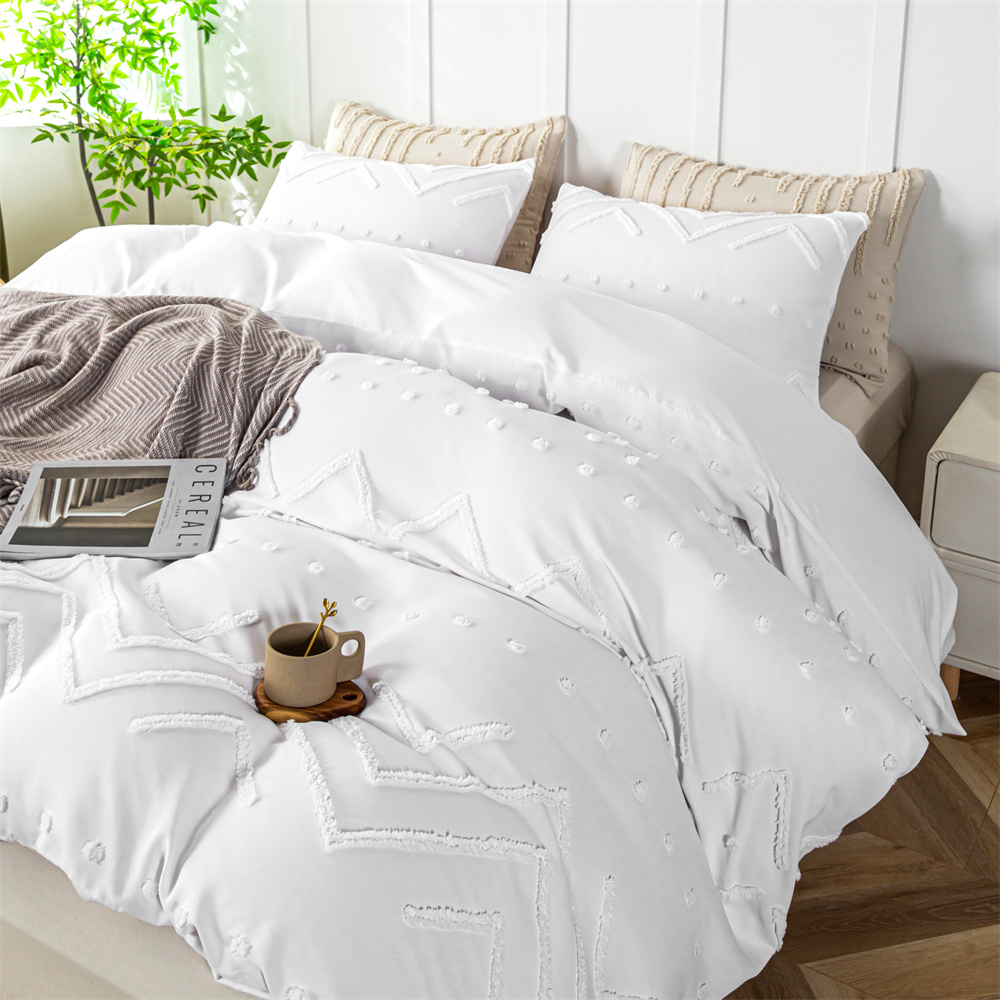 A white queen comforter set with decorative tufts and textured geometric patterns, accompanied by matching pillowcases and a beige throw blanket at the foot of the bed.