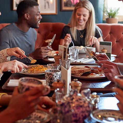 Group of people dining, with various dishes on the table including a pie, and tea served in patterned teapots and cups.
