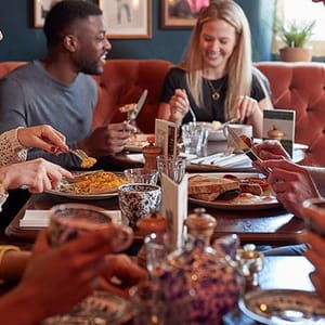 Group of people dining, with various dishes on the table including a pie, and tea served in patterned teapots and cups.