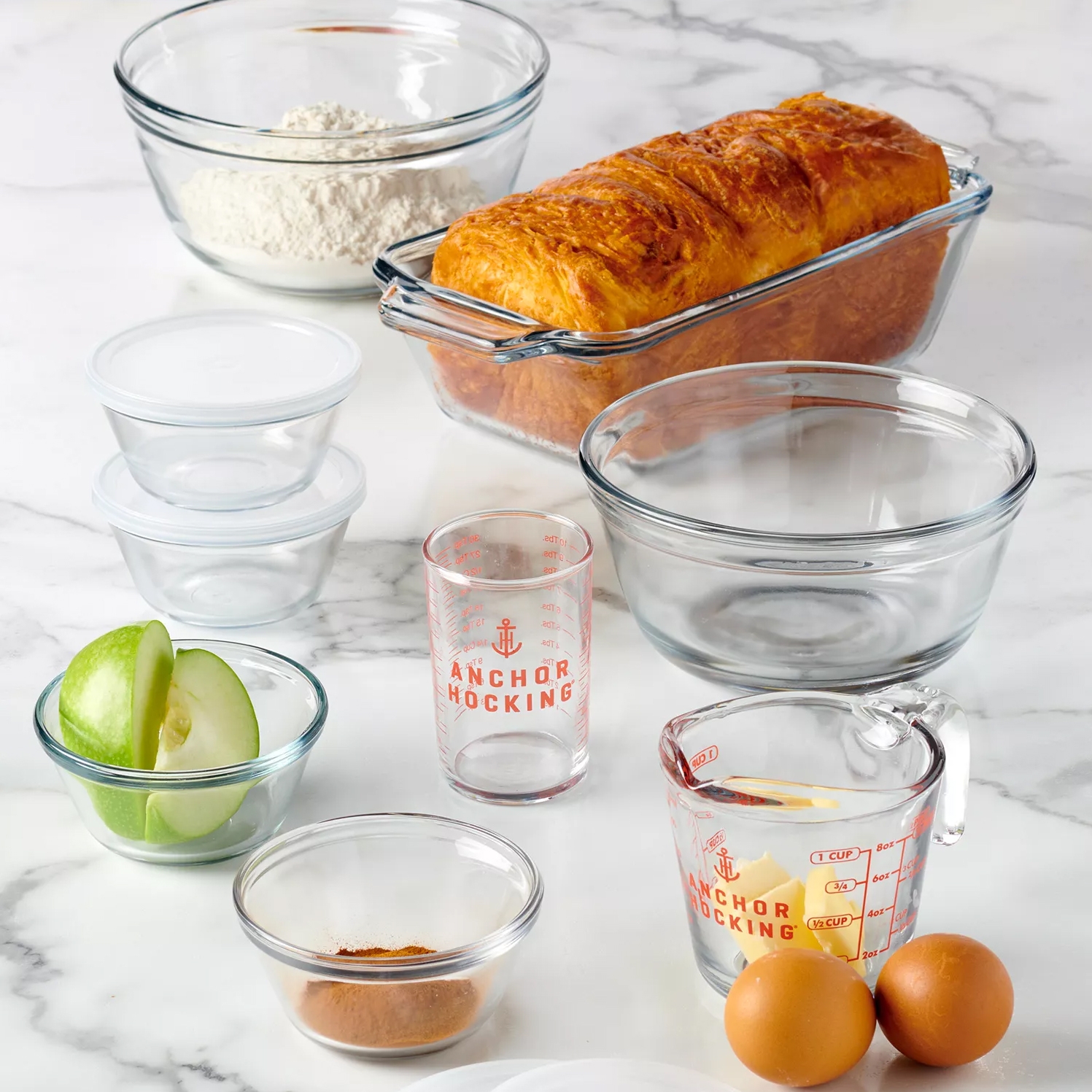 A collection of clear glass kitchenware including bowls, measuring cups, and storage containers, featuring \“Anchor Hocking\“ branding. Also shown are a loaf of bread, two eggs, green apple slices, and cinnamon powder.