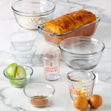 A collection of clear glass kitchenware including bowls, measuring cups, and storage containers, featuring \“Anchor Hocking\“ branding. Also shown are a loaf of bread, two eggs, green apple slices, and cinnamon powder.