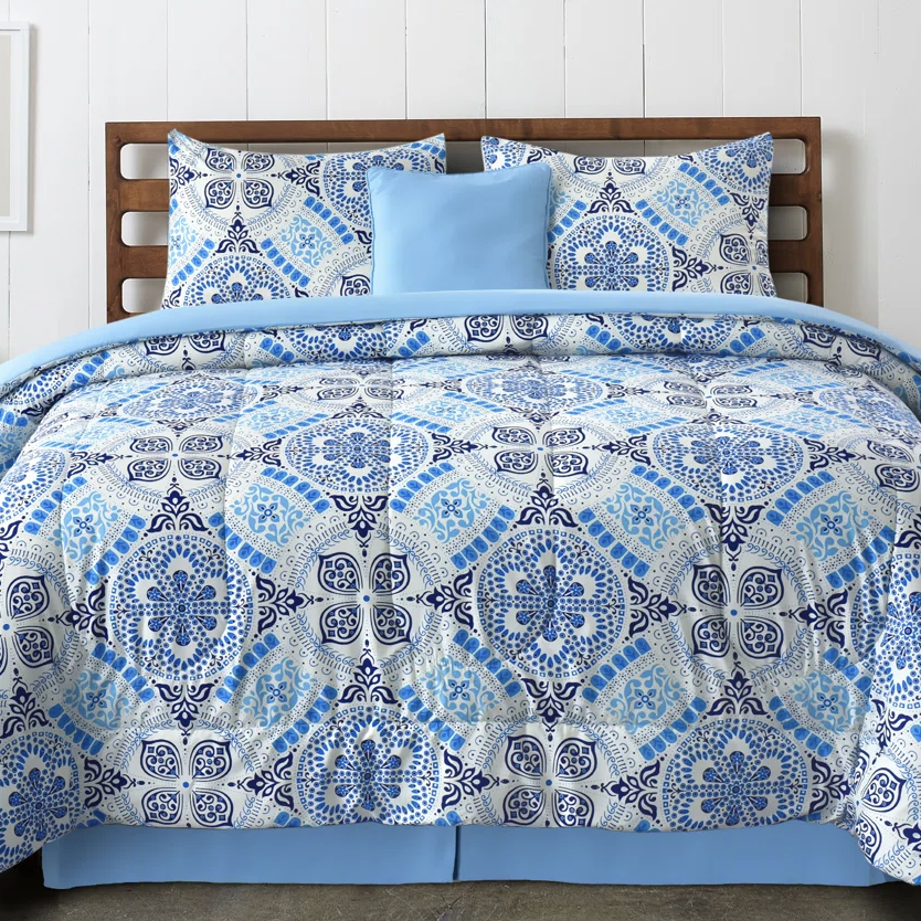 A bed is dressed with a blue and white patterned comforter set, featuring intricate geometric designs, accompanied by matching pillow shams and a solid light blue accent pillow.