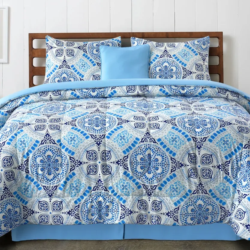 A bed is dressed with a blue and white patterned comforter set, featuring intricate geometric designs, accompanied by matching pillow shams and a solid light blue accent pillow.