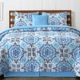 A bed is dressed with a blue and white patterned comforter set, featuring intricate geometric designs, accompanied by matching pillow shams and a solid light blue accent pillow.