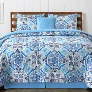 A bed is dressed with a blue and white patterned comforter set, featuring intricate geometric designs, accompanied by matching pillow shams and a solid light blue accent pillow.