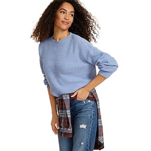 A woman is wearing a light blue sweater with a plaid shirt tied around her waist and distressed blue jeans.