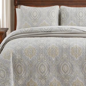 A quilt set featuring a light gray and beige paisley pattern design, including a quilt and two matching pillow shams, displayed on a wooden bed frame.