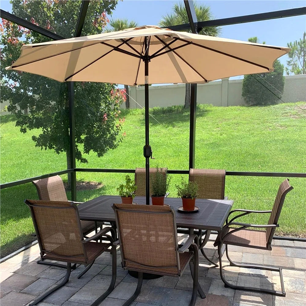 A 9' crank tilt patio umbrella with a beige canopy provides shade over an outdoor dining set, which includes a rectangular table and six brown mesh chairs on a tiled patio. Potted plants rest on the table with a view of a grassy yard beyond.