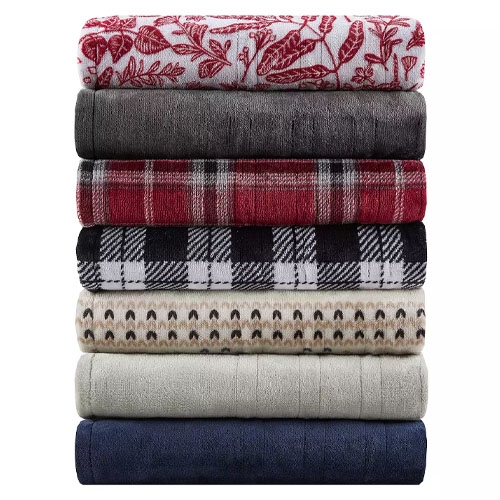 A stack of seven plush fleece blankets in various colors and patterns, including floral, solid gray, red plaid, black and white checkered, and geometric designs.
