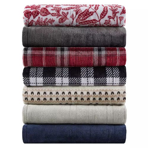 A stack of seven plush fleece blankets in various colors and patterns, including floral, solid gray, red plaid, black and white checkered, and geometric designs.
