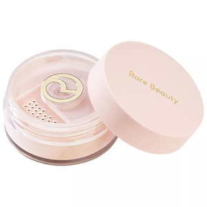 A round container with a pink lid labeled \“Rare Beauty\“ holds a translucent loose setting powder, featuring a logo-shaped sifter.