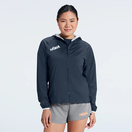 A woman is wearing a dark full-zip hooded jacket with the HOKA logo on the chest and a contrasting light grey band along the hem.