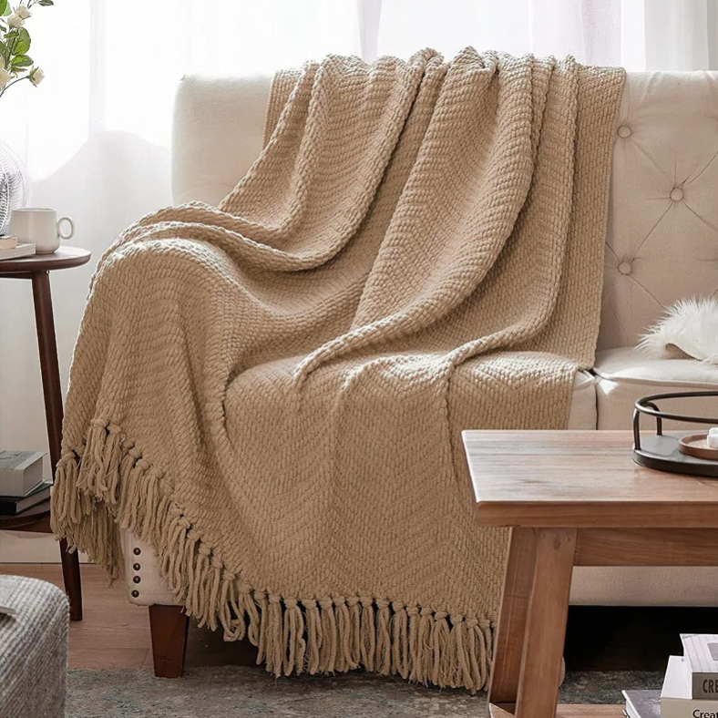 A beige, textured knit throw blanket with fringe edges is draped over a sofa, accompanied by a wooden coffee table with a decorative tray, a side table with books and a coffee cup, and floor-length curtains.