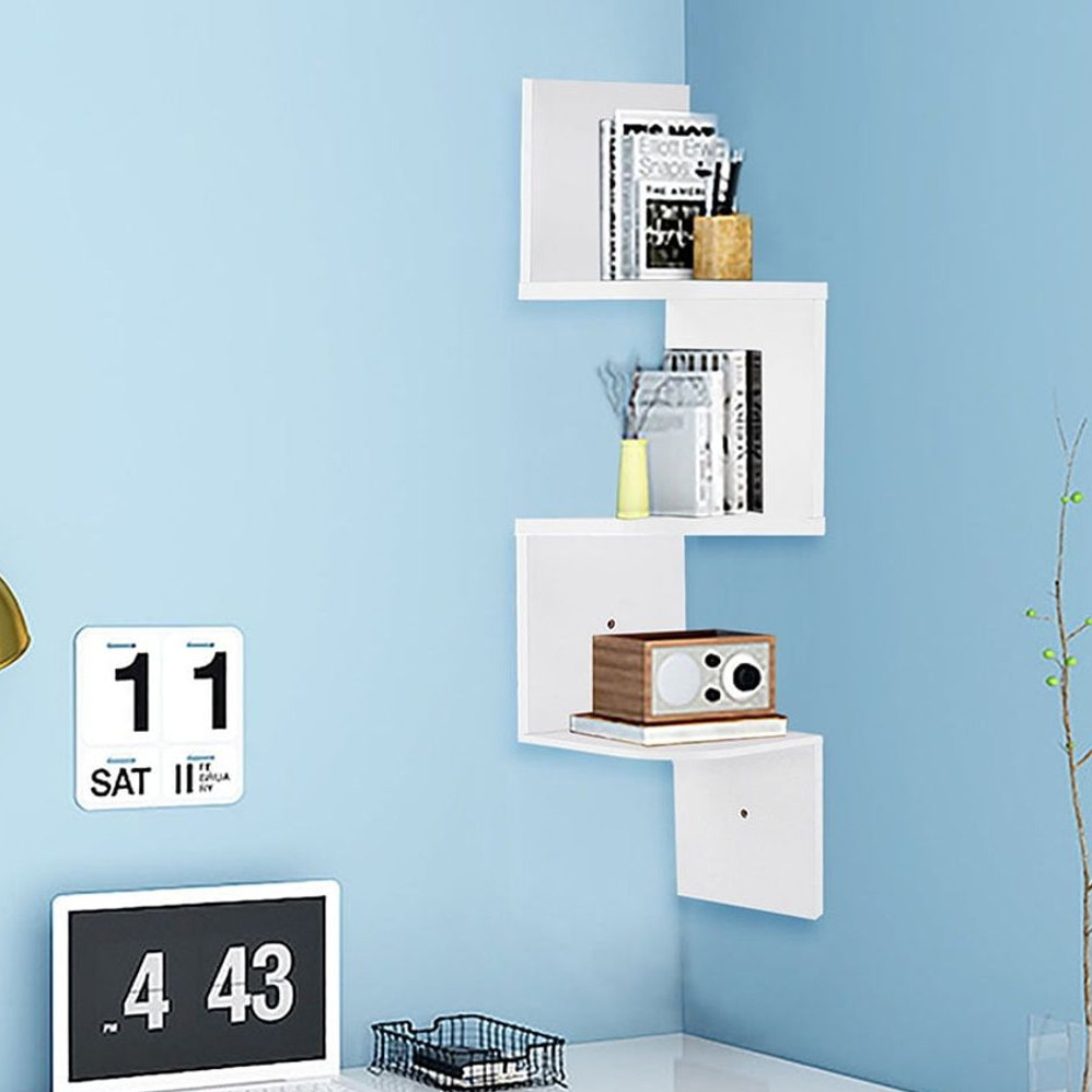 White corner wall shelf against a blue wall holds books, small decor, and a wooden radio. Below, a small black and white digital clock and a wire basket on a desk are visible.