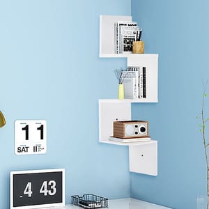White corner wall shelf against a blue wall holds books, small decor, and a wooden radio. Below, a small black and white digital clock and a wire basket on a desk are visible.