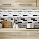 Modern kitchen features a black and white mosaic tile backsplash with wooden cabinets. A stainless steel pot, wooden cutting boards, whisks, and a potted plant are on the countertop.