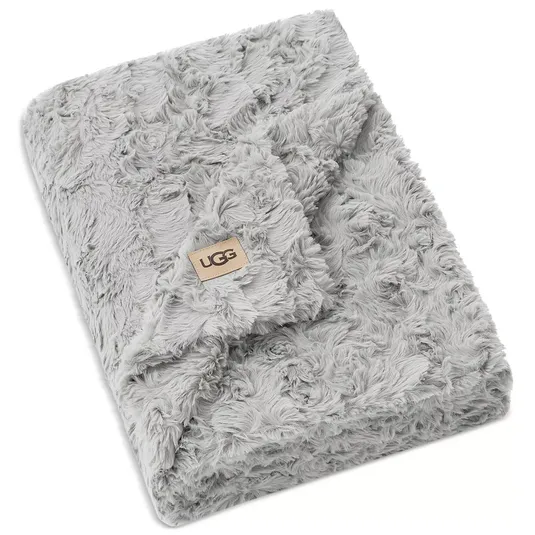 A soft, grey faux fur blanket with the UGG logo on a tan patch, featuring a textured, plush design.