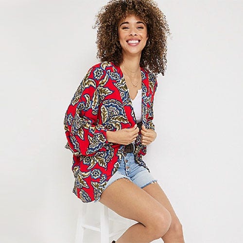 Woman wearing red printed blazer and denim shorts, smiling and sitting on a stool.