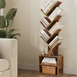 A wooden bookshelf with a unique zigzag design holds various books, alongside a square base featuring additional books and a storage box. A small diffuser is placed on top, next to a potted plant.