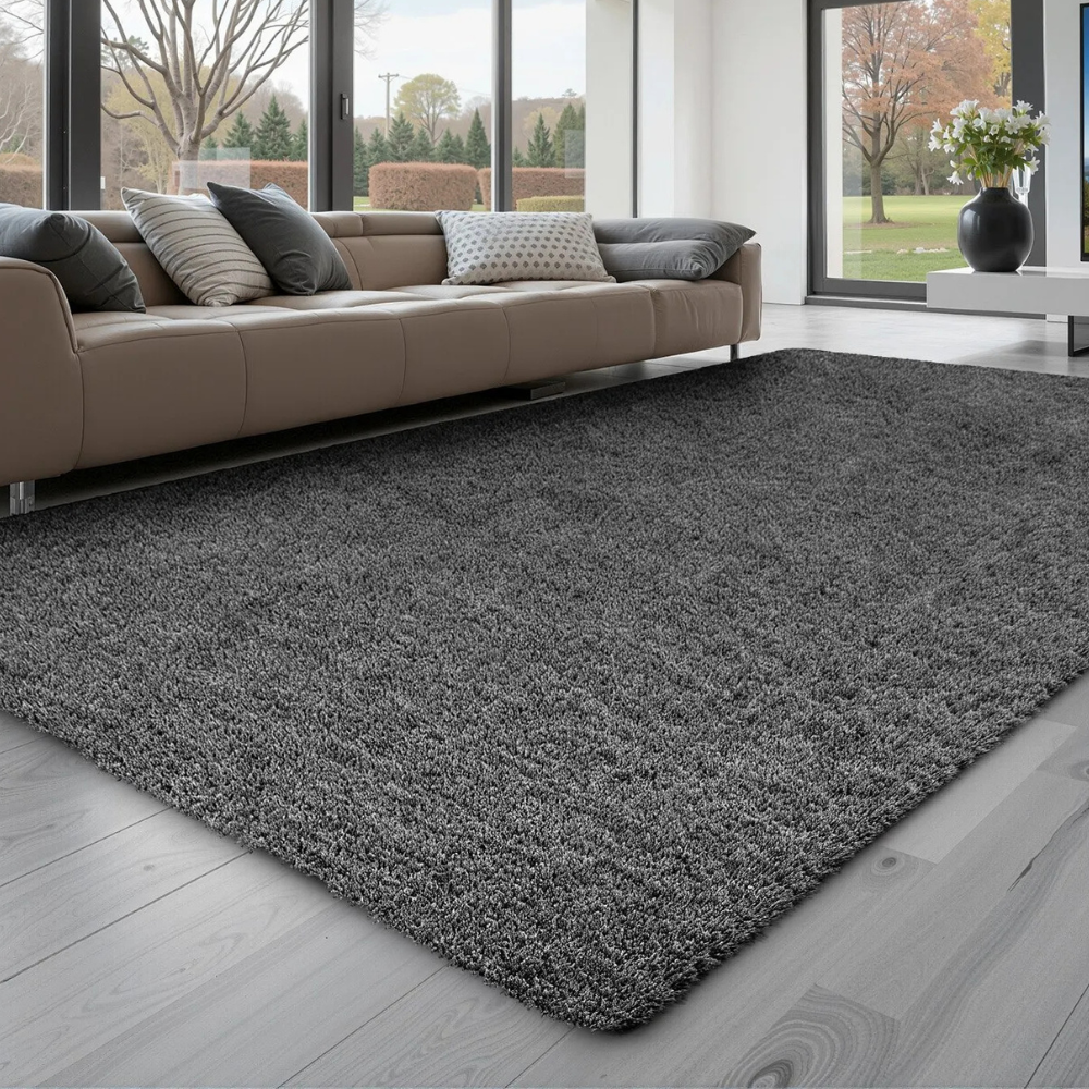 A cozy gray shag rug lies on a light wooden floor in front of a modern beige sectional sofa with various gray pillows, set in a spacious living room with large windows.