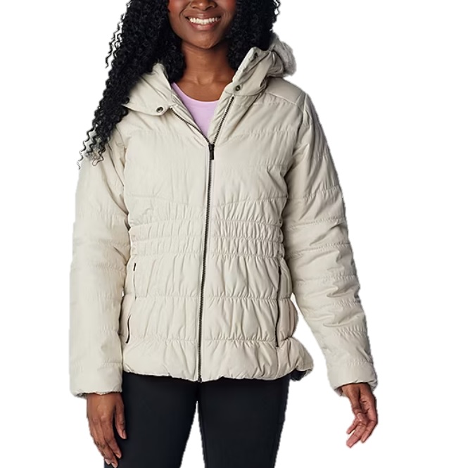 A woman wears a Columbia quilted jacket in a light color. The jacket features a hood, front zipper, and gathered elastic waist, providing a snug fit.