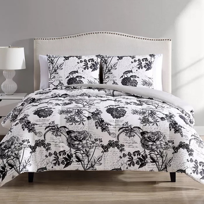 The image shows a bed with a black and white floral-patterned comforter and pillow shams. A white headboard and a bedside table with a lamp are also visible.