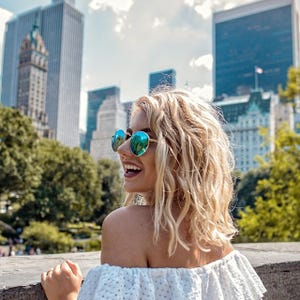 A woman with blonde hair is wearing reflective sunglasses and an off-the-shoulder white top, standing in a city park with skyscrapers in the background.