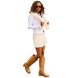 A person in a park wears a white long-sleeve top, cream-colored shorts, a light sweater draped over the shoulders, and tan knee-high boots.