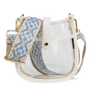 A clear handbag with white accents and a wide, adjustable shoulder strap featuring a blue and beige geometric pattern, adorned with gold-tone hardware.