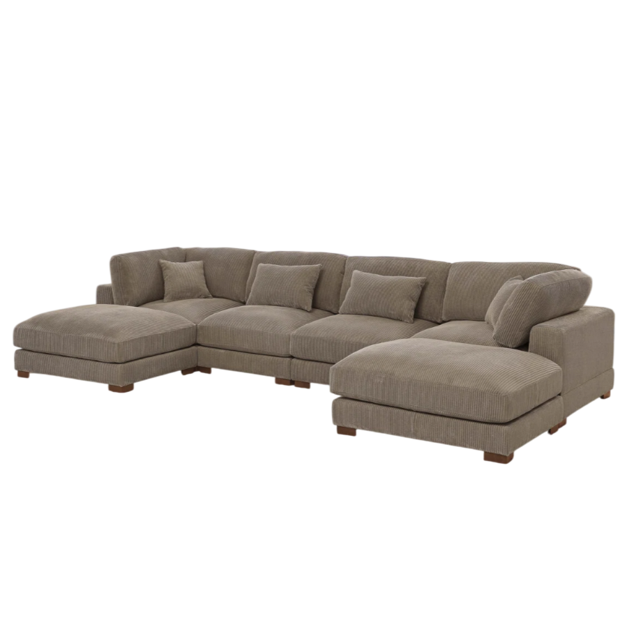 Large L-shaped sectional sofa in brown corduroy with multiple cushions, placed on a light-colored textured rug in a spacious, well-lit living room with large windows, a chandelier, and a bookshelf.