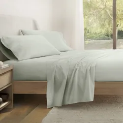 A neatly made bed with light-colored sheets and pillowcases sits next to a wooden nightstand with a lamp and books. A large window showcases greenery outside.