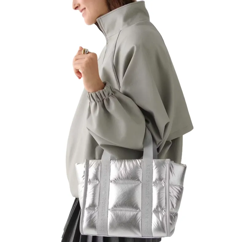 A person is carrying a metallic silver Kate Spade tote bag with quilted texture, featuring the brand name on the straps, while wearing a light-colored high-collared jacket.