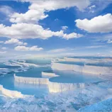 Terraced mineral pools filled with clear blue water, against a bright blue sky with fluffy white clouds.