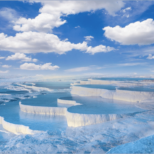 Terraced mineral pools filled with clear blue water, against a bright blue sky with fluffy white clouds.
