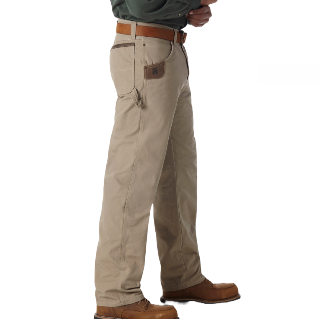 Wrangler Men's Carpenter Pants in beige feature a relaxed fit with multiple pockets, a hammer loop, and contrast stitching. They pair with a brown belt and boots, suitable for work or casual wear.